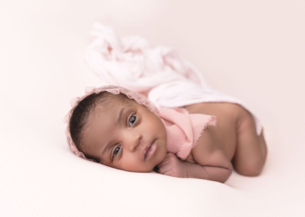 A newborn baby dressed in pink resting on a soft surface, looking at the camera with a calm expression.