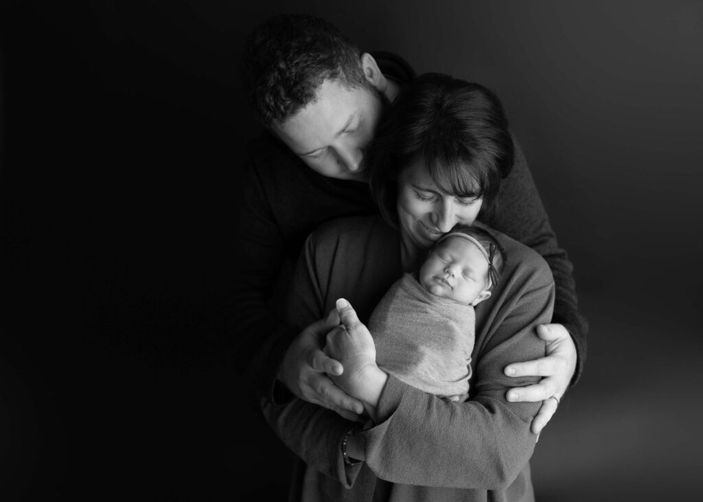 A black and white photo of a man and woman cradling and embracing a sleeping newborn.